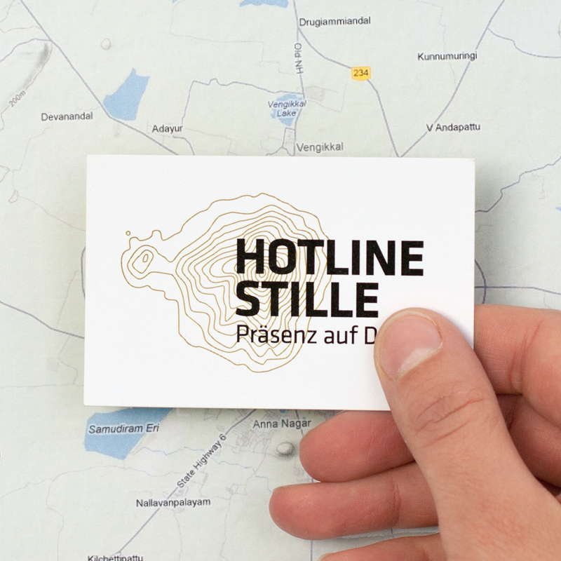Eine Hand hält die Visitenkarte mit dem Logo von Hotline Stille vor eine Landkarte. Das Logo besteht aus dem Schriftzug und gelben Höhenlinien.