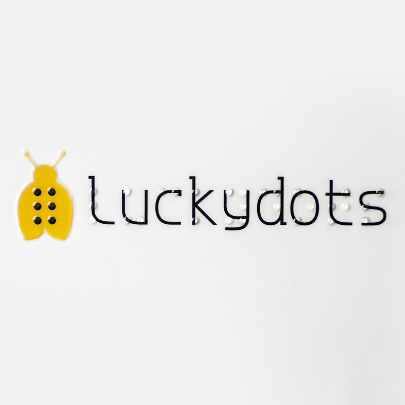 Das Logo von LUCKY DOTS besteht aus einem gelben Marienkäfer. Über den Schriftzeichen sind Braillepunkte zu sehen